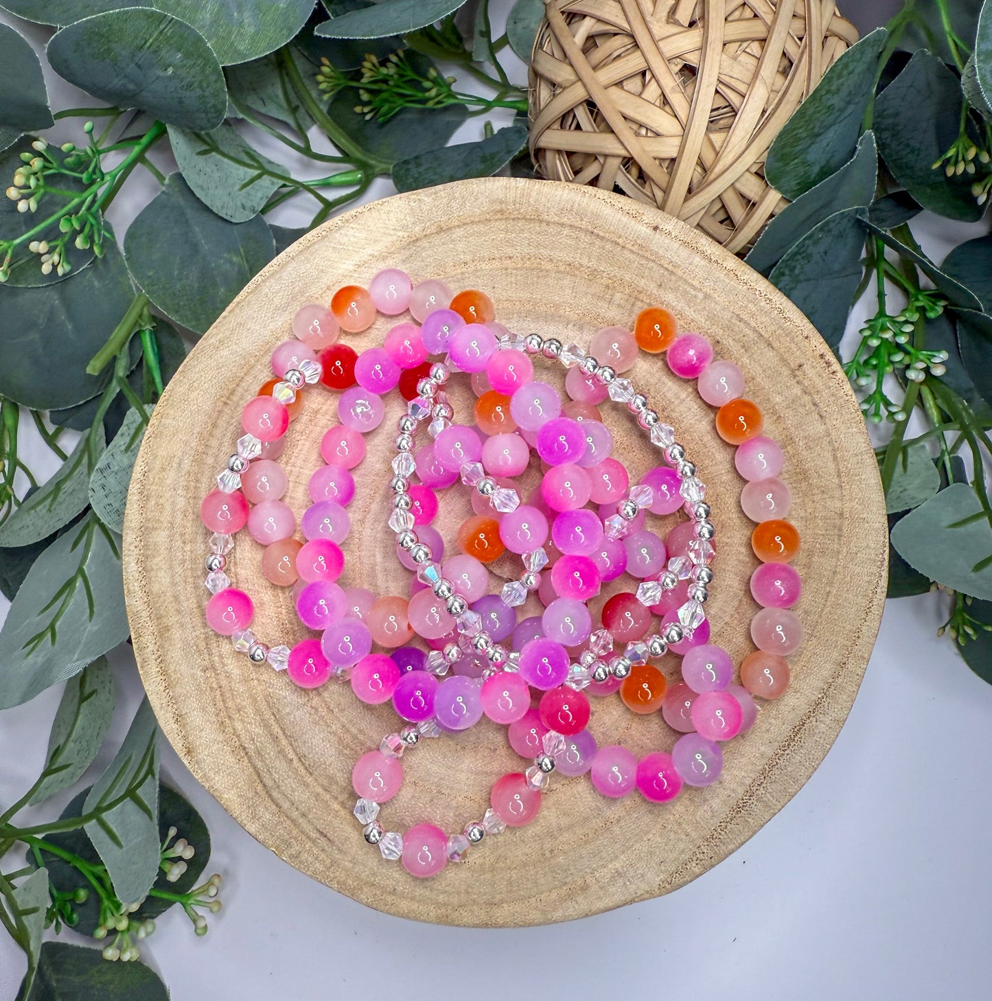 A wooden plate holds a collection of colorful beaded bracelets, with a wicker basket and greenery in the background.