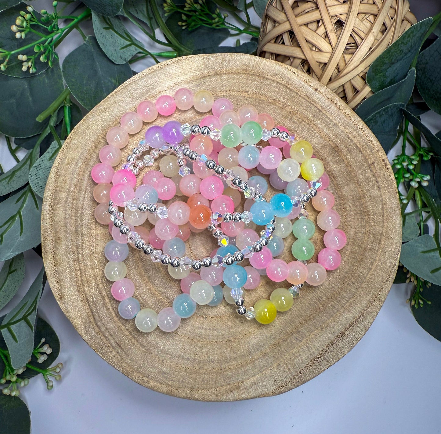 A collection of colorful beaded bracelets and necklaces are arranged on a wooden surface, surrounded by greenery.