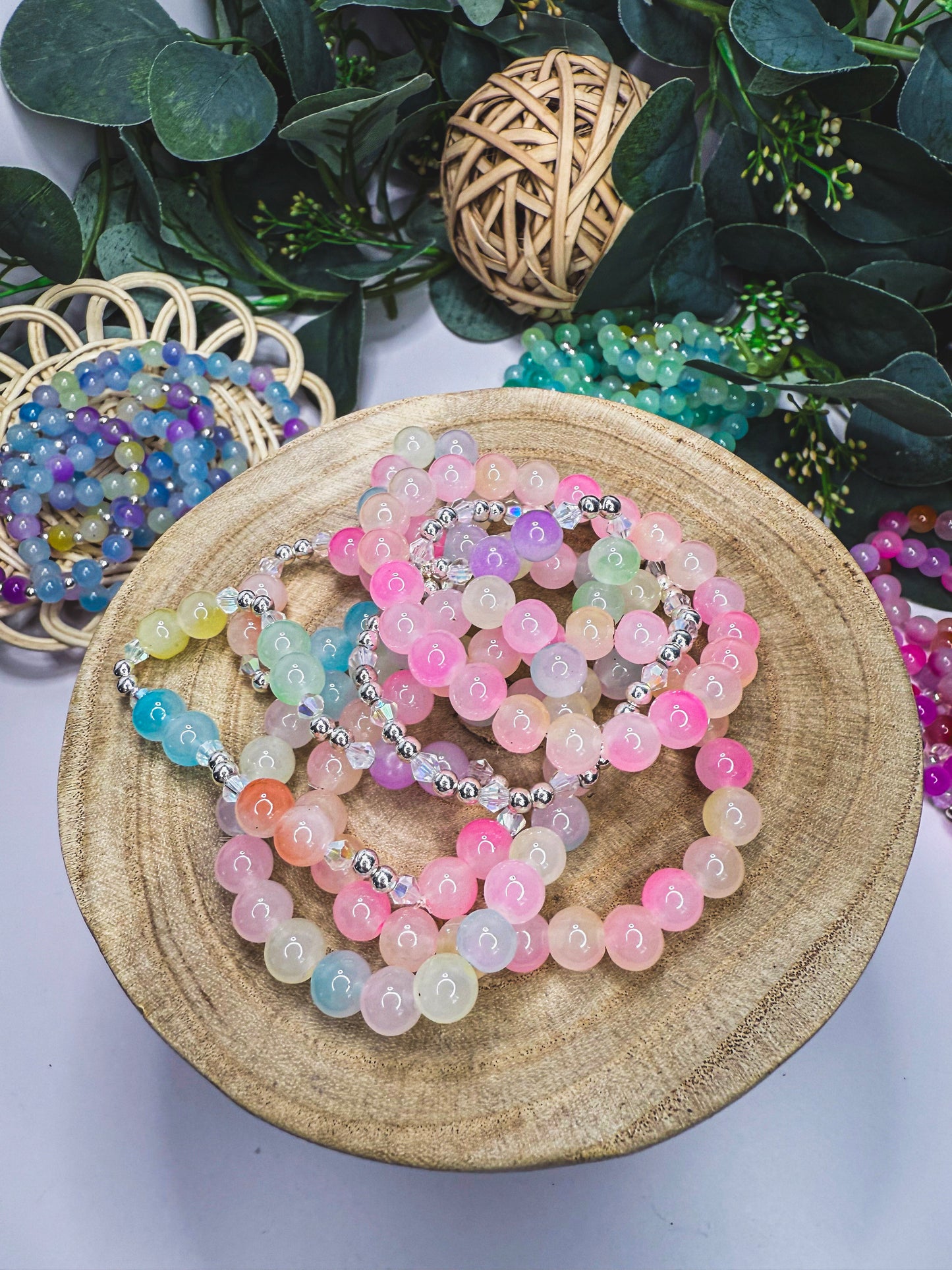 A wooden bowl filled with multicolored beaded bracelets is placed on a white surface, surrounded by greenery and a woven basket.