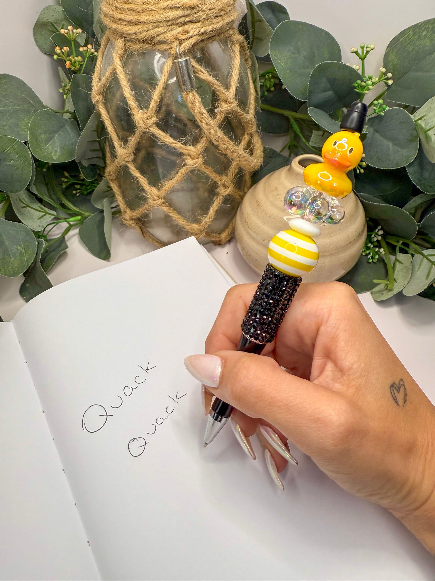 A hand holding a pen with a quack quack design, writing on a notebook, with a small decorative item and greenery in the background.