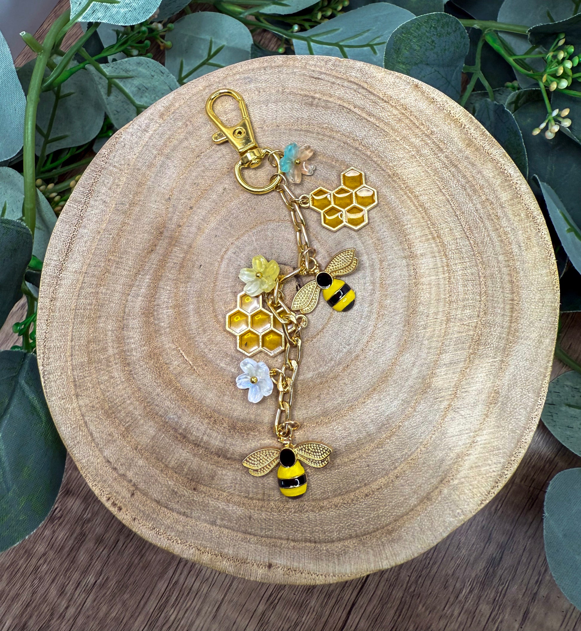 A wooden plate with a decorative keychain featuring a bee and honeycomb design, surrounded by greenery.