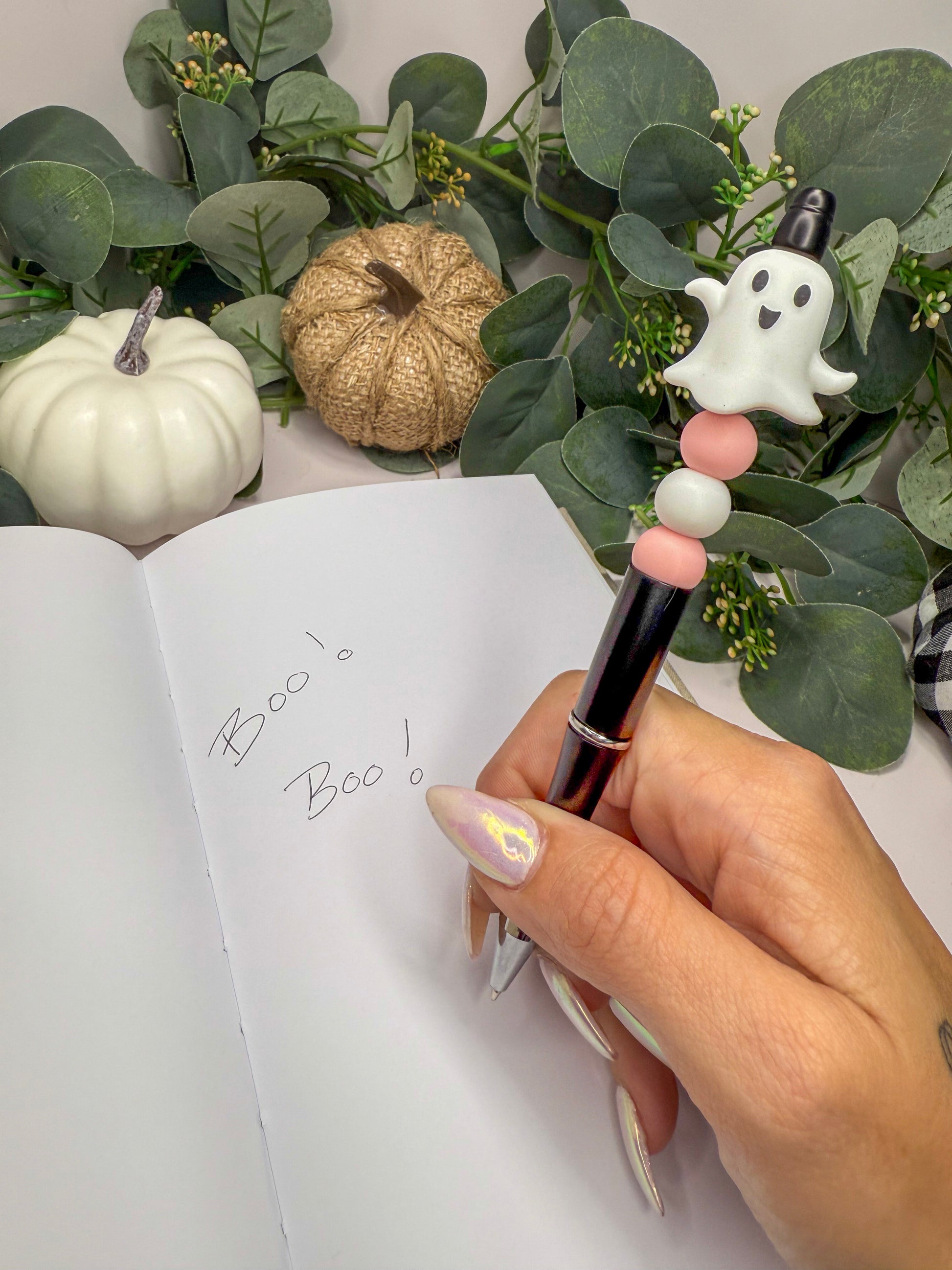 A hand holding a pen with a ghost-like design, writing on a notebook, surrounded by Halloween decorations such as a pumpkin and greenery.