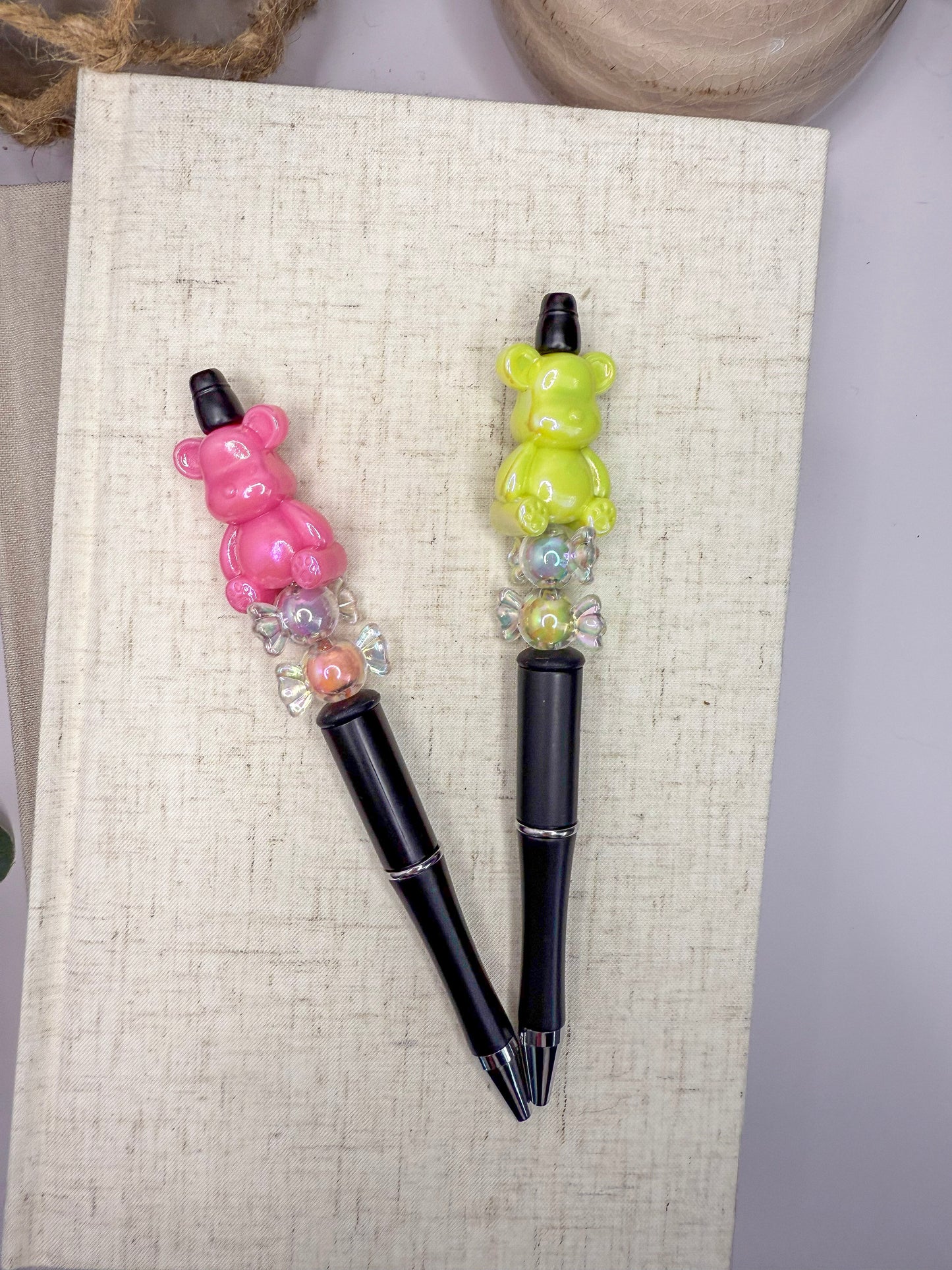 Two colorful pens with bear-shaped handles are placed on a white surface, with a beige book and a wooden object in the background.