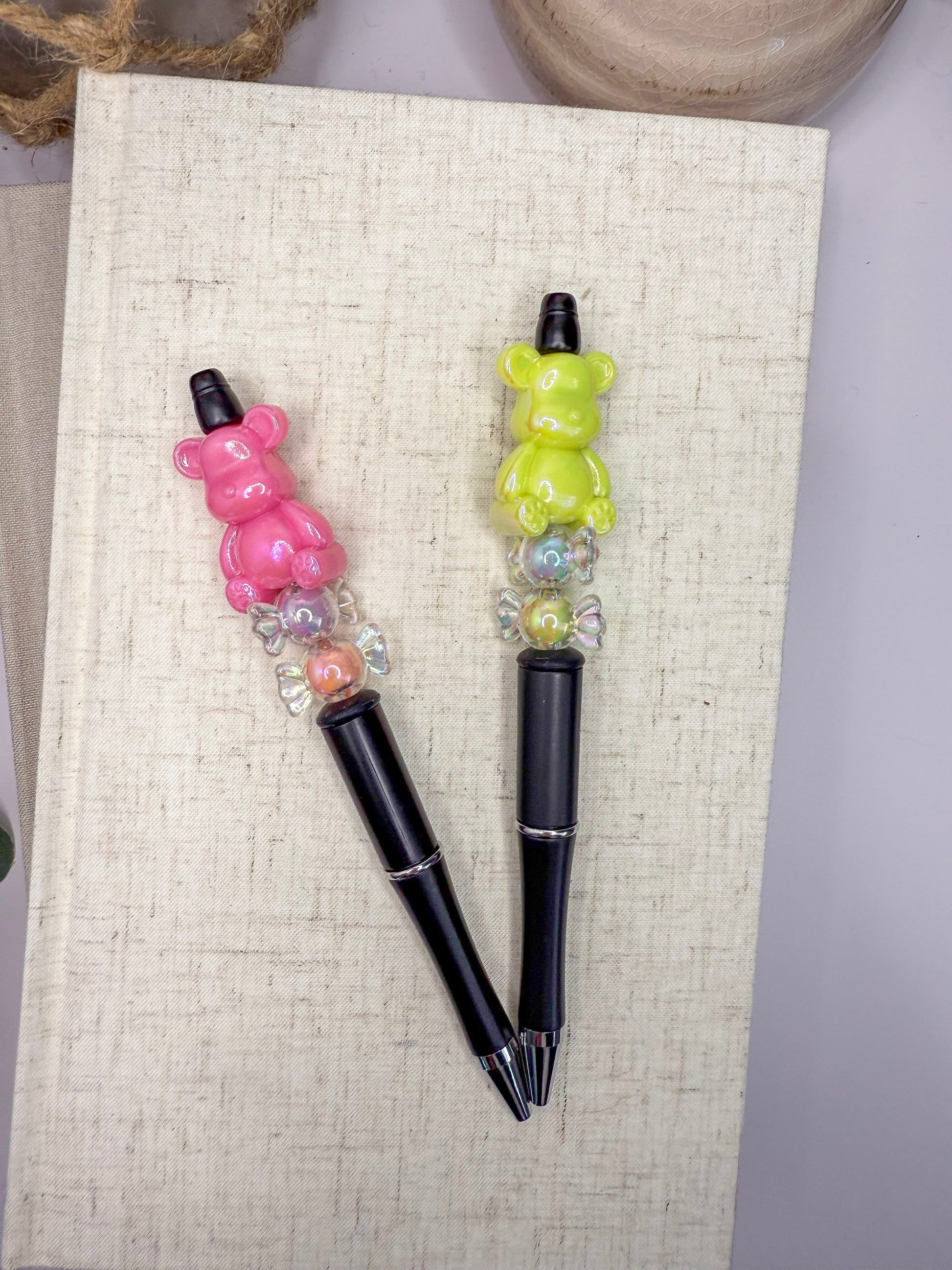 Two colorful pens with bear-shaped handles are placed on a white surface, with a beige book and a wooden object in the background.