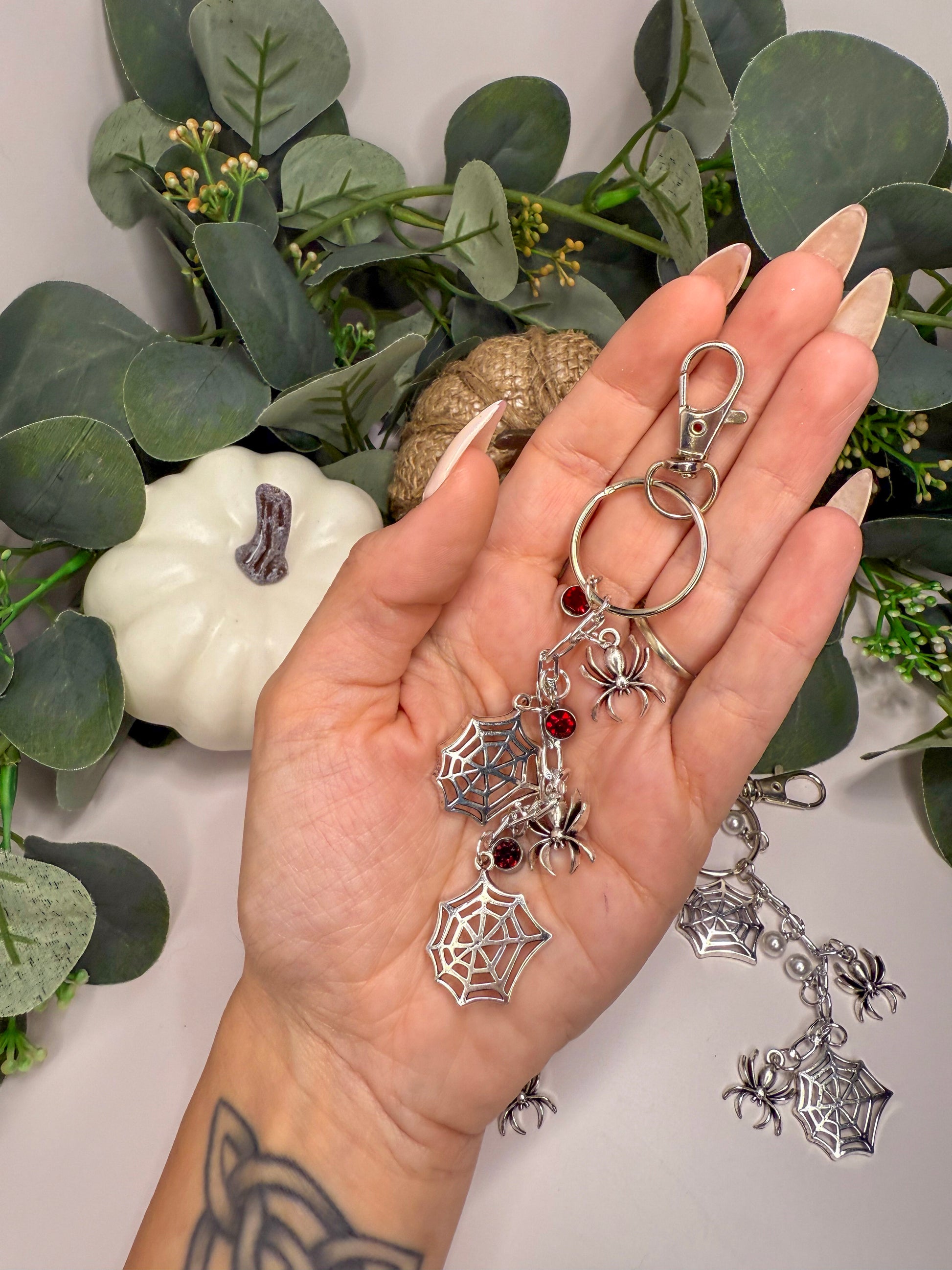 A hand holding a keychain with spider charm and a white pumpkin decoration in the background.