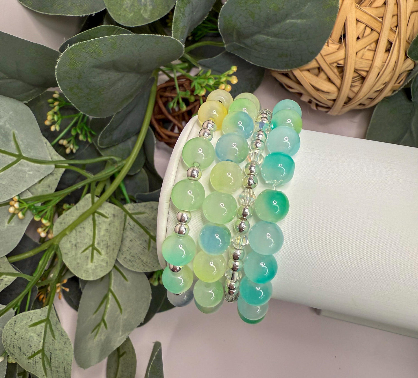 A beaded bracelet with green and blue stones is displayed on a white surface, surrounded by greenery and a woven basket.