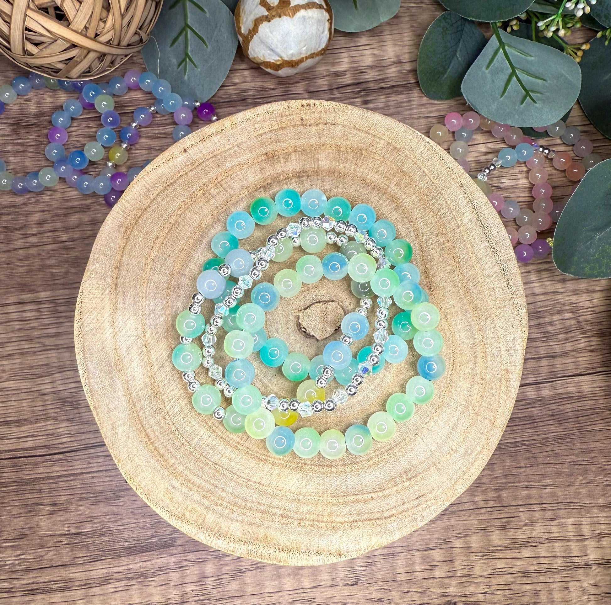 A collection of colorful beaded bracelets and necklaces arranged on a wooden surface, with some greenery visible in the background.