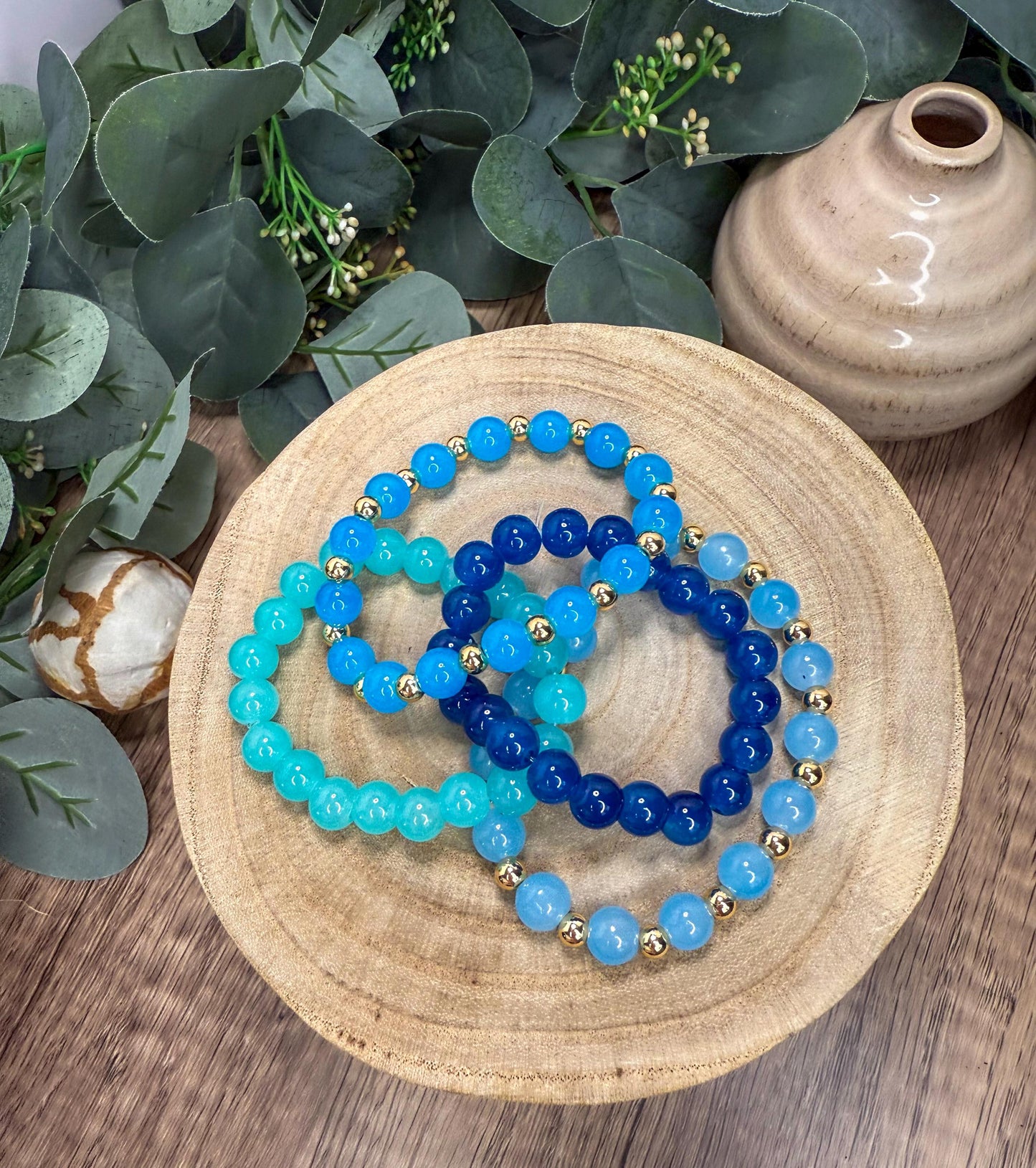 A wooden plate holds a collection of colorful beaded bracelets, with blue and blue-green beads arranged in a circular pattern. The plate is placed on a wooden surface, with a vase and some greenery in the background.