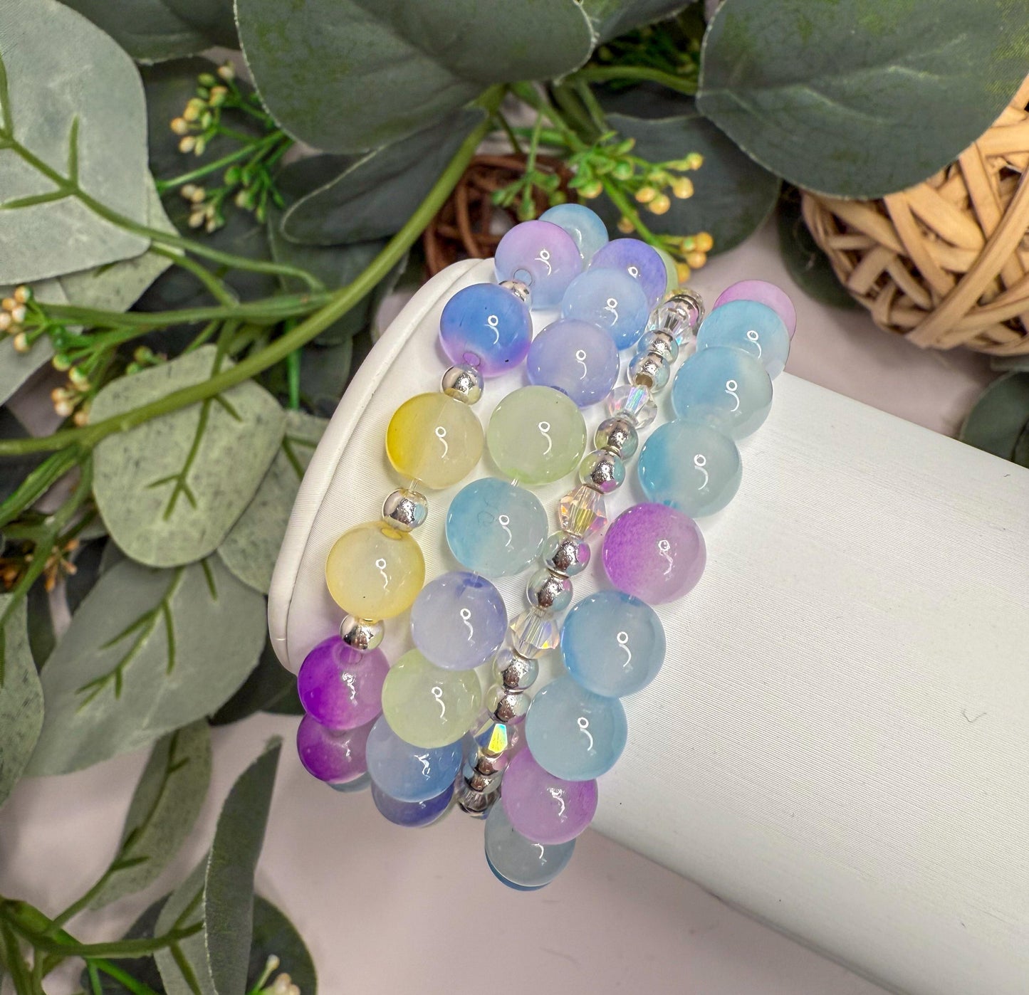 A colorful beaded bracelet is displayed on a white stand, surrounded by greenery and flowers.
