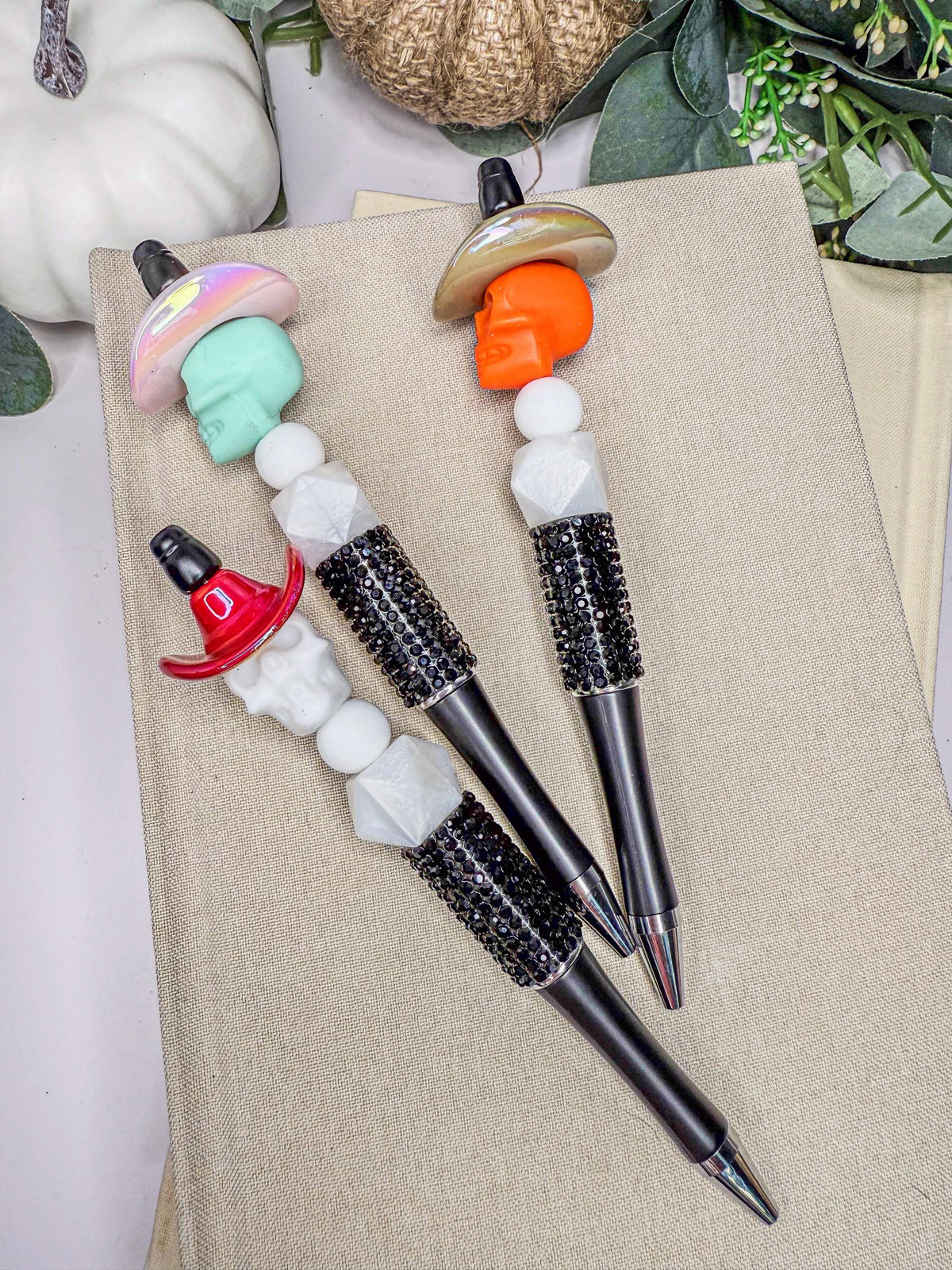 three pens with unique designs, including skull and mushroom heads, placed on a beige surface.