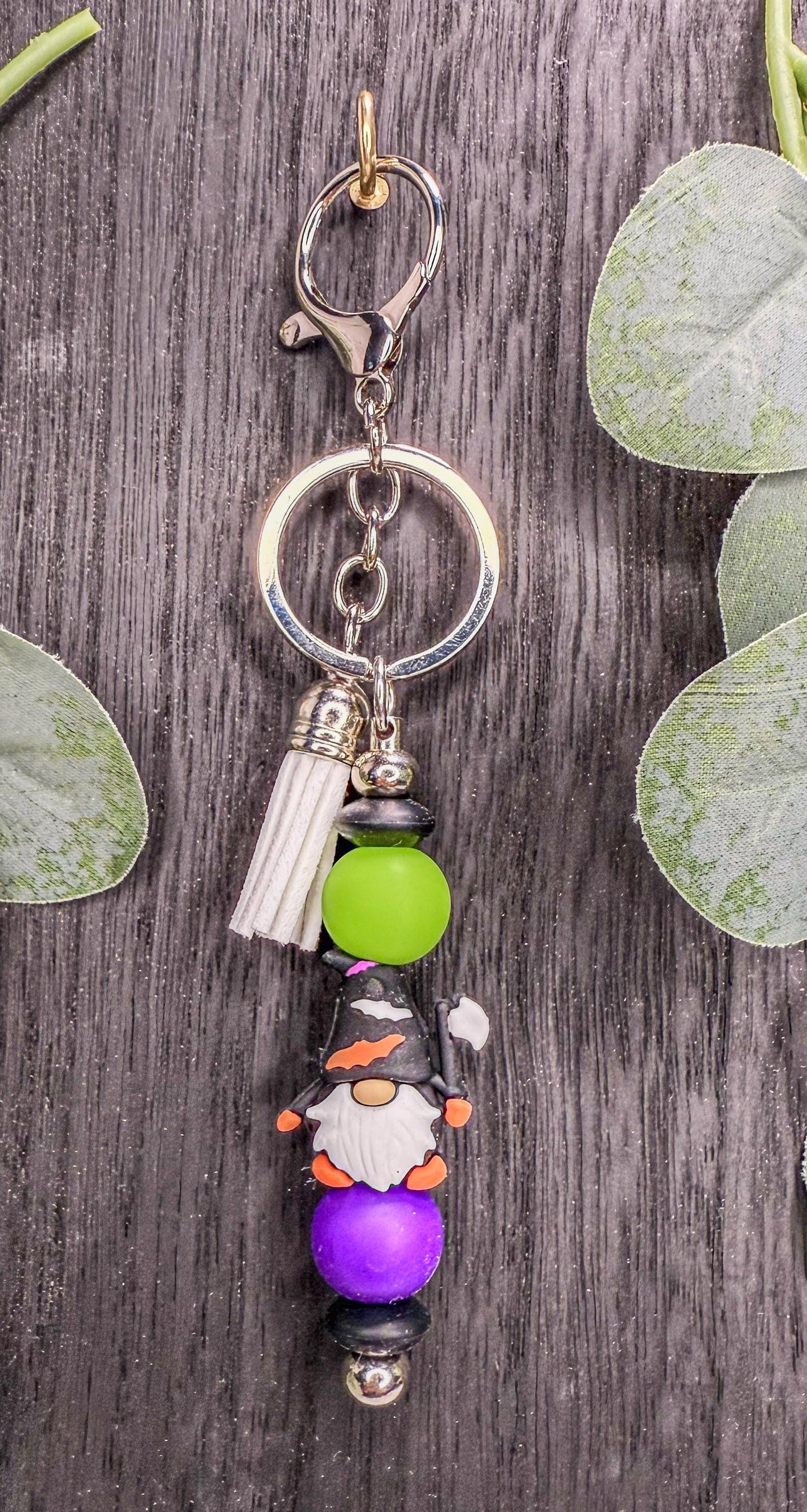 A colorful keychain with a gnome design, featuring green beads and purple beads, is displayed on a wooden surface with some leaves.