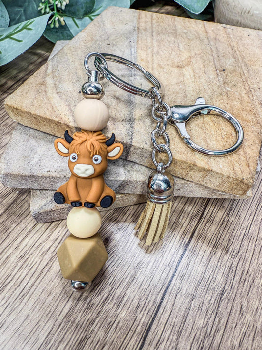 A keychain featuring a cartoon cow character with horns and a tassel, sitting on a beige bead.