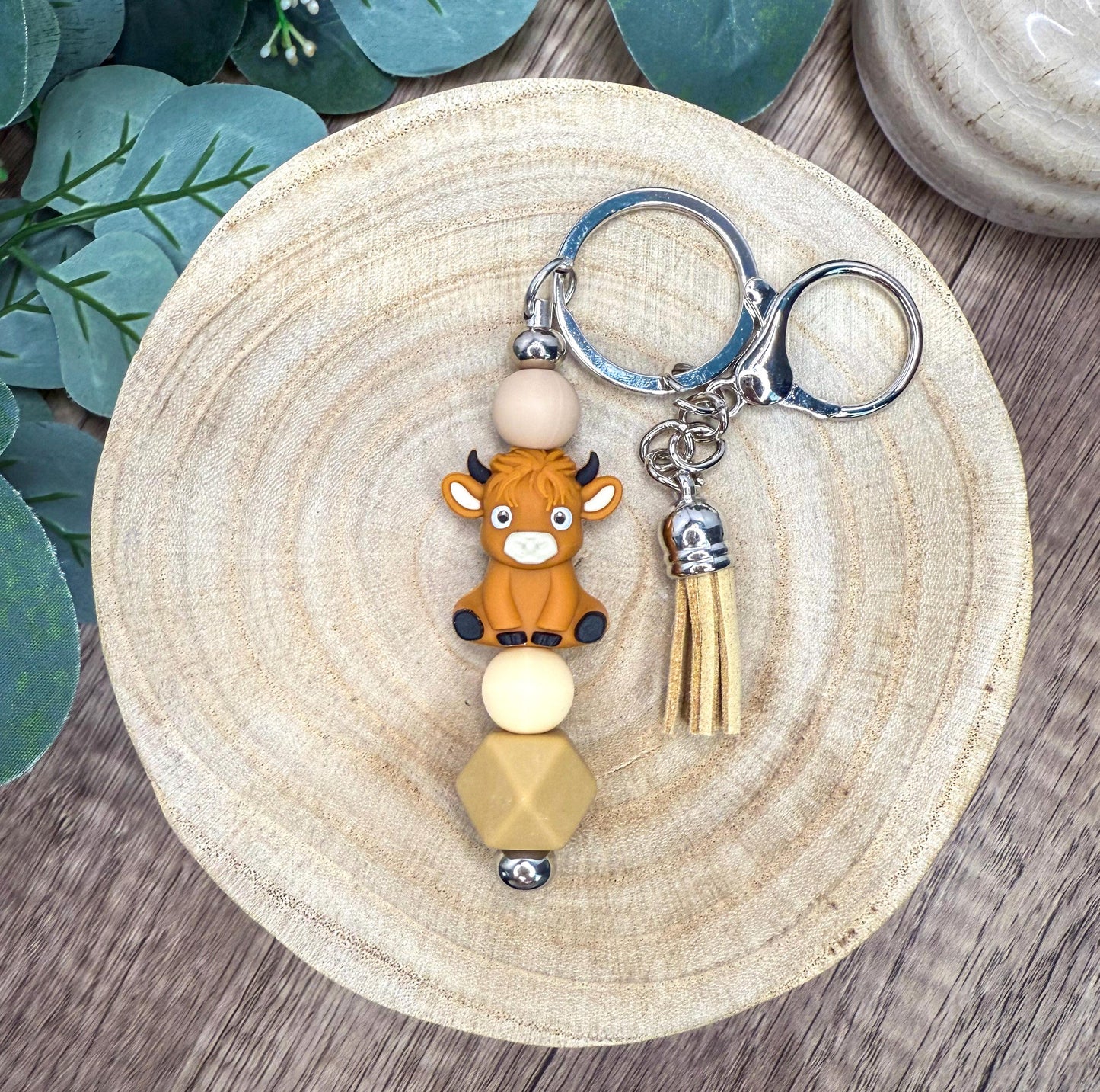 A keychain featuring a small brown cow figure with horns and a tassel, placed on a wooden surface with some greenery in the background.