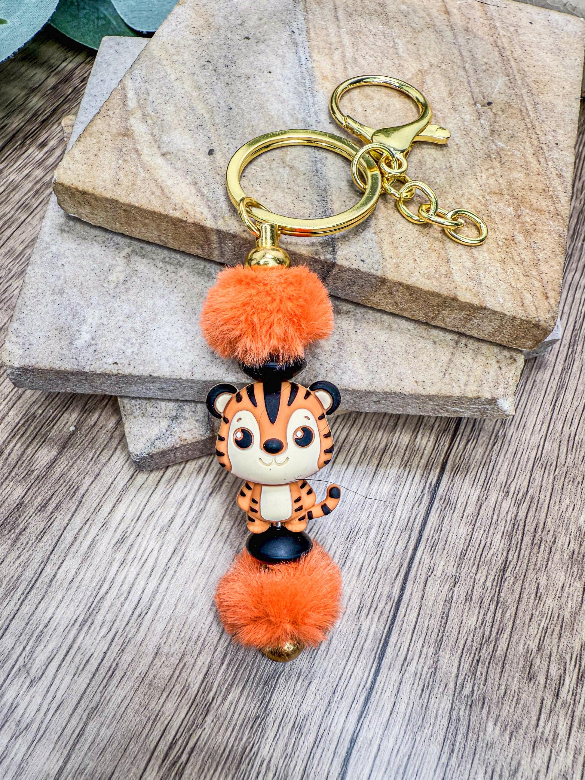 A keychain featuring a small tiger figurine with orange pom poms, placed on a wooden surface.