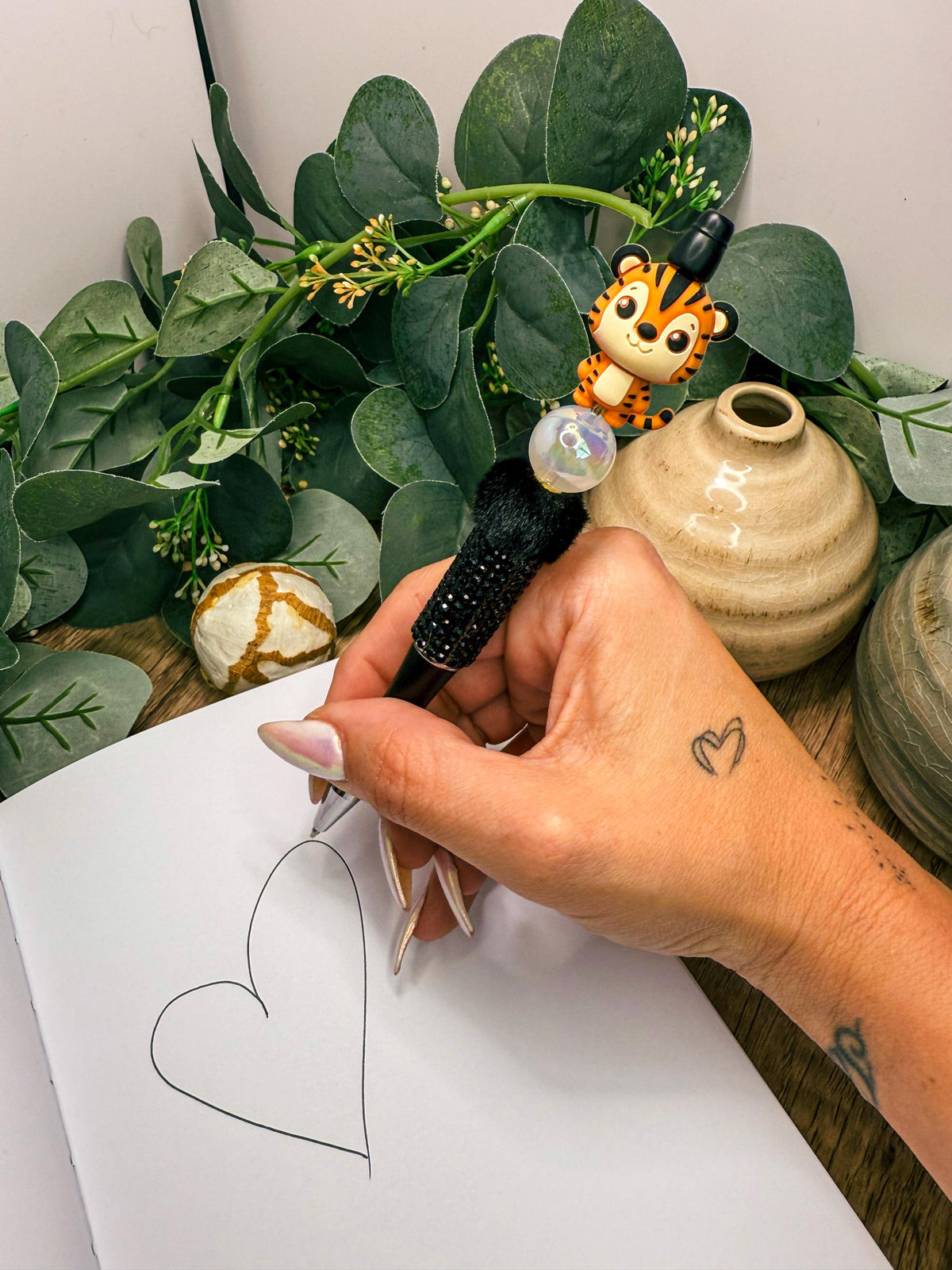 A hand is holding a pen and drawing a heart shape on a piece of paper, with a small tiger figurine and a plant in the background.