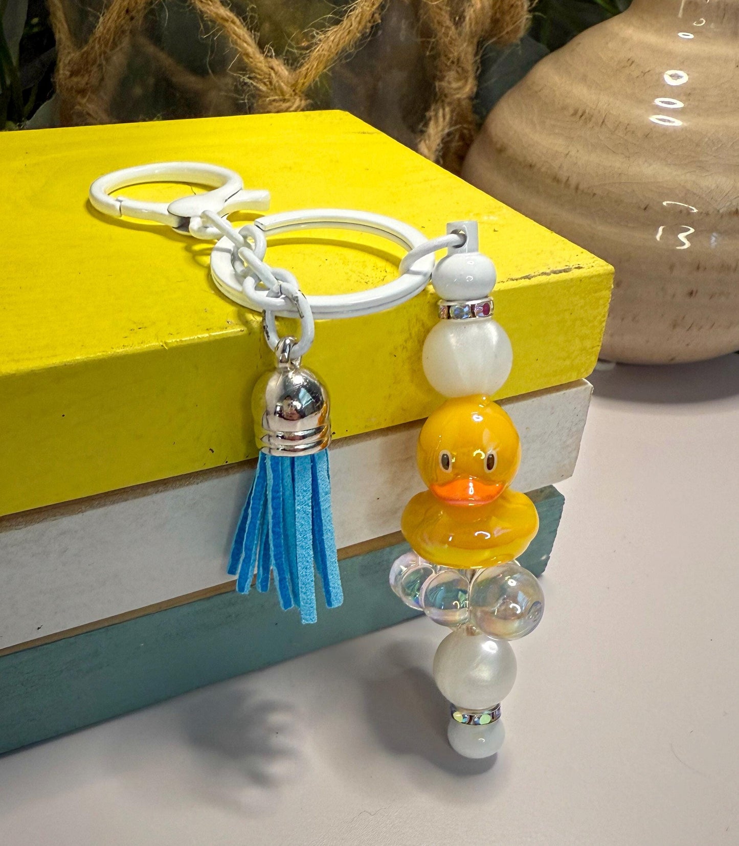 A keychain with a yellow duck charm and a blue tassel is displayed on top of a stack of books.