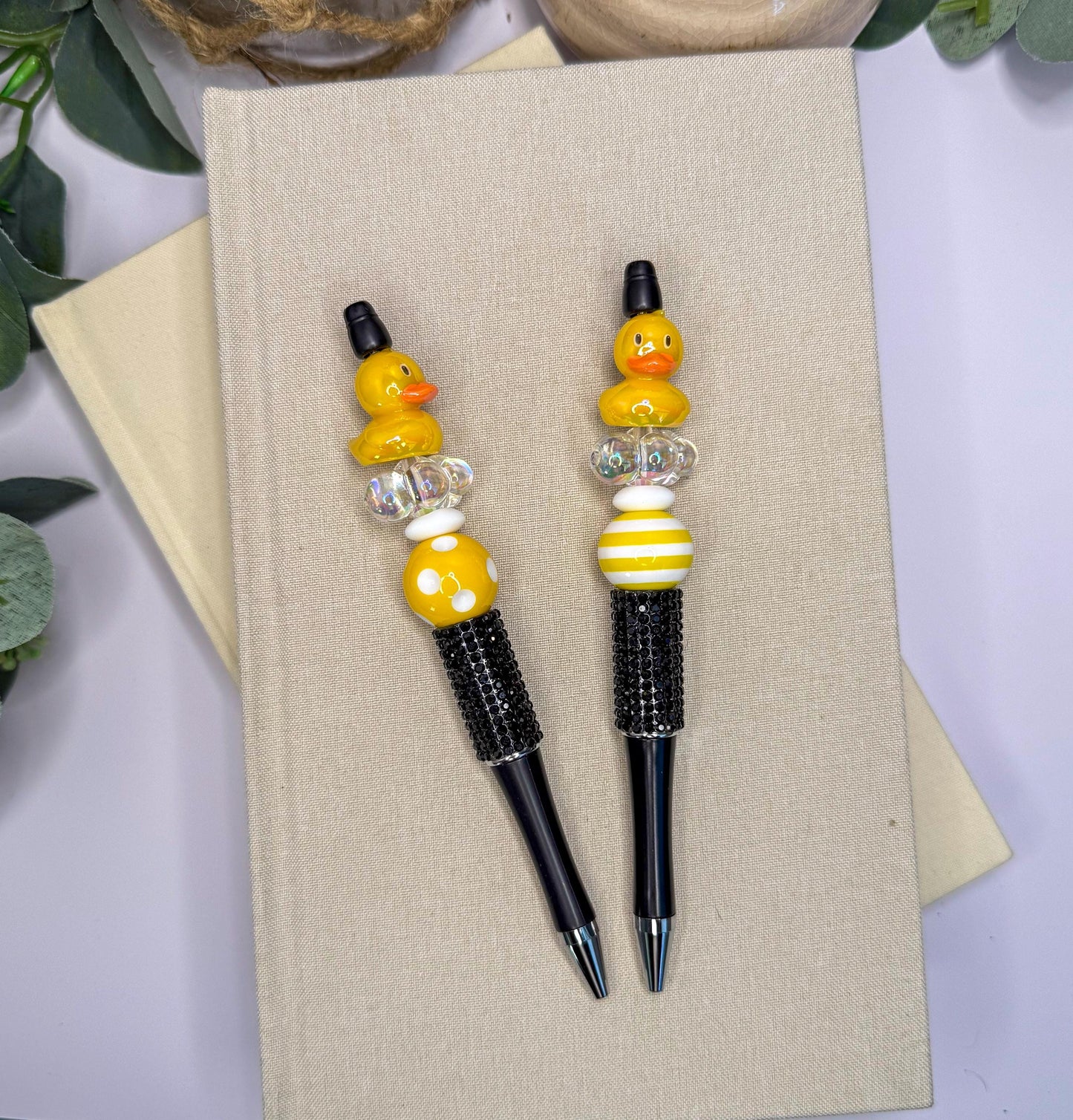 two yellow and black pens with decorative elements, such as beads and a duck, placed on top of a beige surface.
