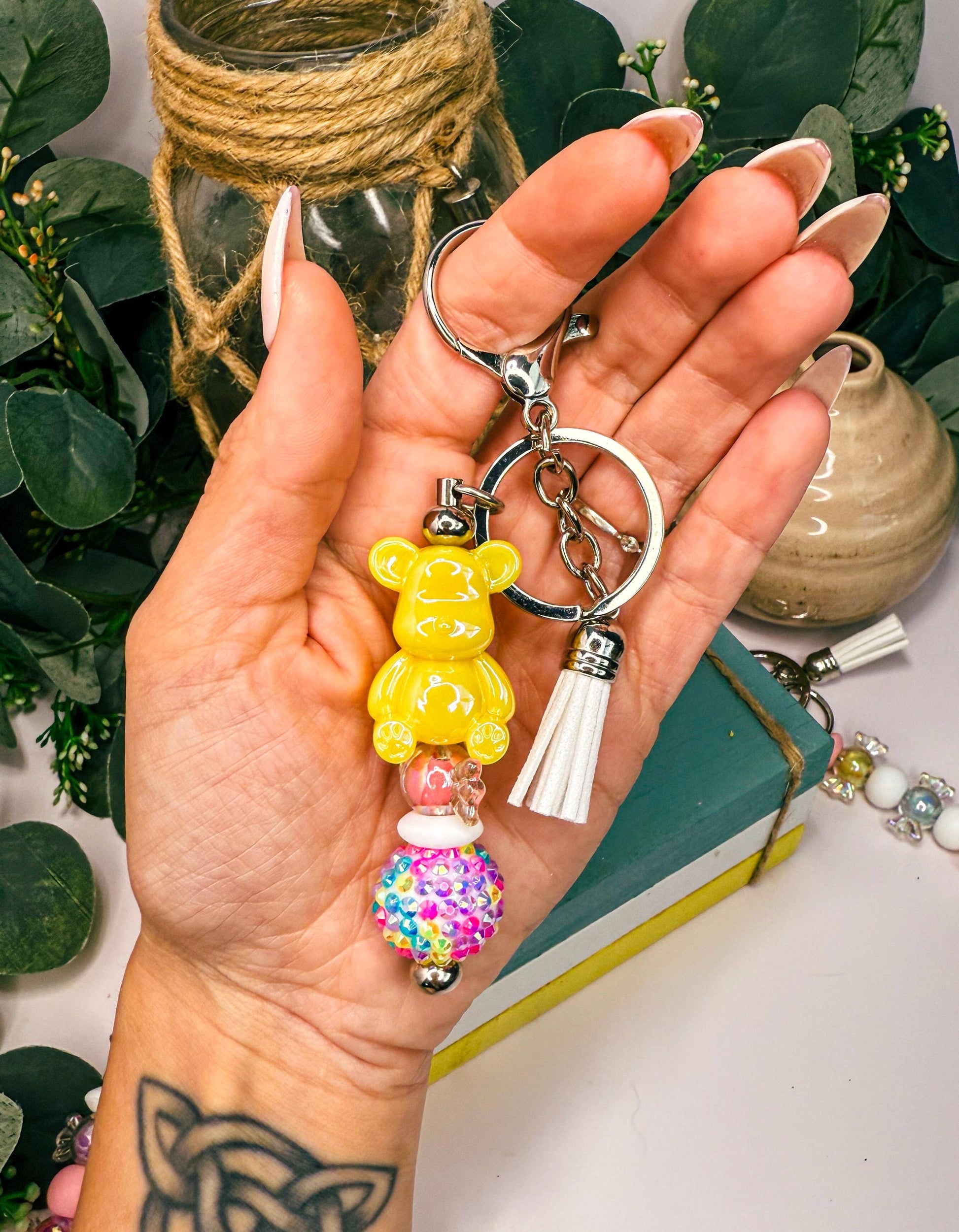 A hand holding a keychain with a yellow teddy bear charm and a colorful gumball charm.