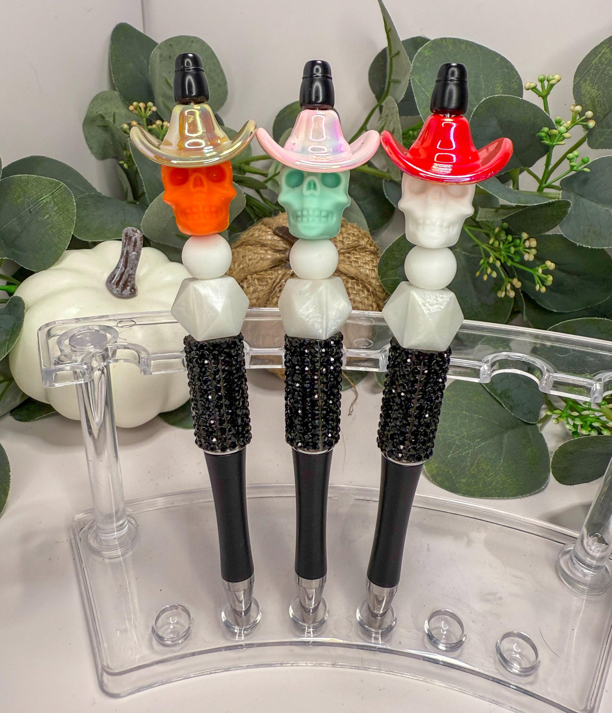 Three colorful pens with unique designs, including a skull, a hat, and a flower, are displayed on a clear stand.