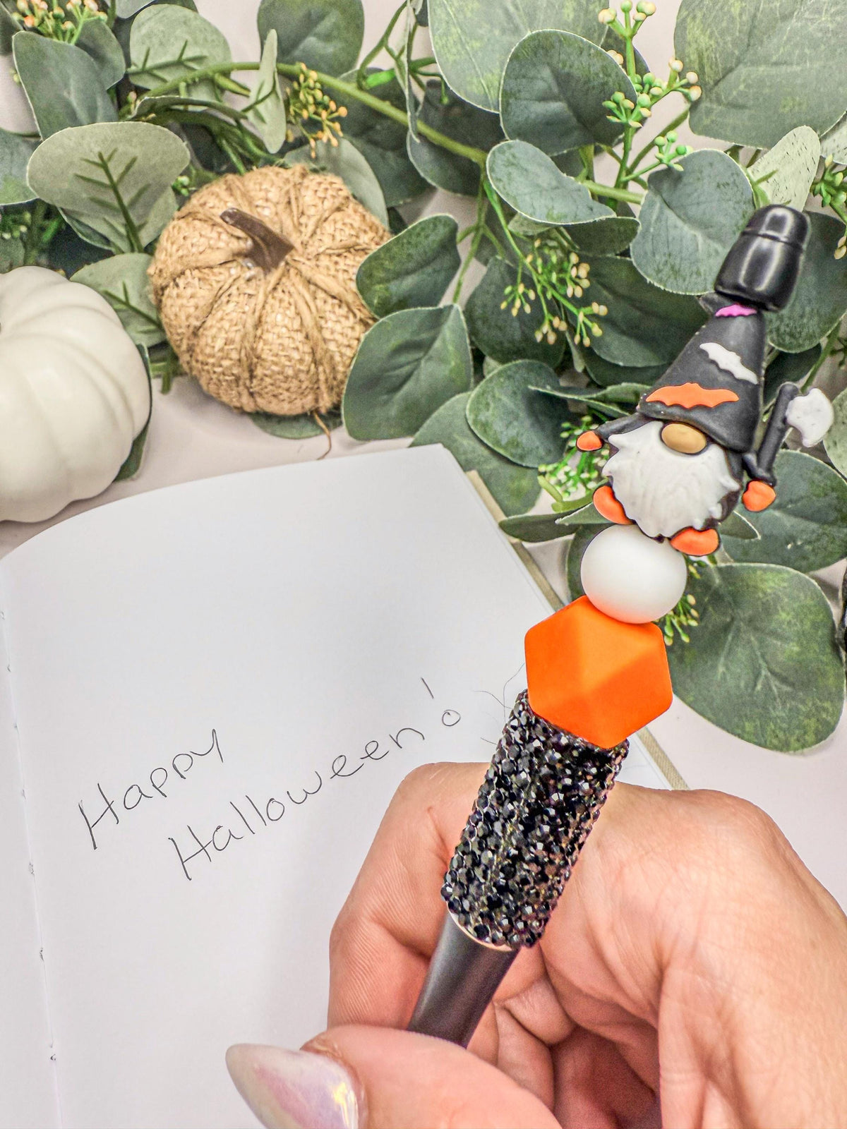 A hand holding a pen with a glittery, black and orange design, writing on a piece of paper with a "Happy Halloween" message.