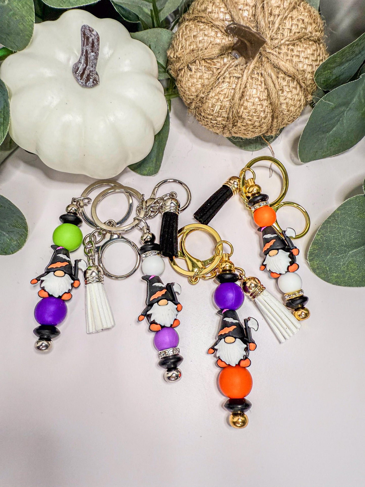 a collection of colorful keychains with gnome-like designs, including purple, orange, and white beads, as well as tassels and charms. The keychains are arranged on a white surface, with a small white pumpkin and a burlap pumpkin visible in the background.