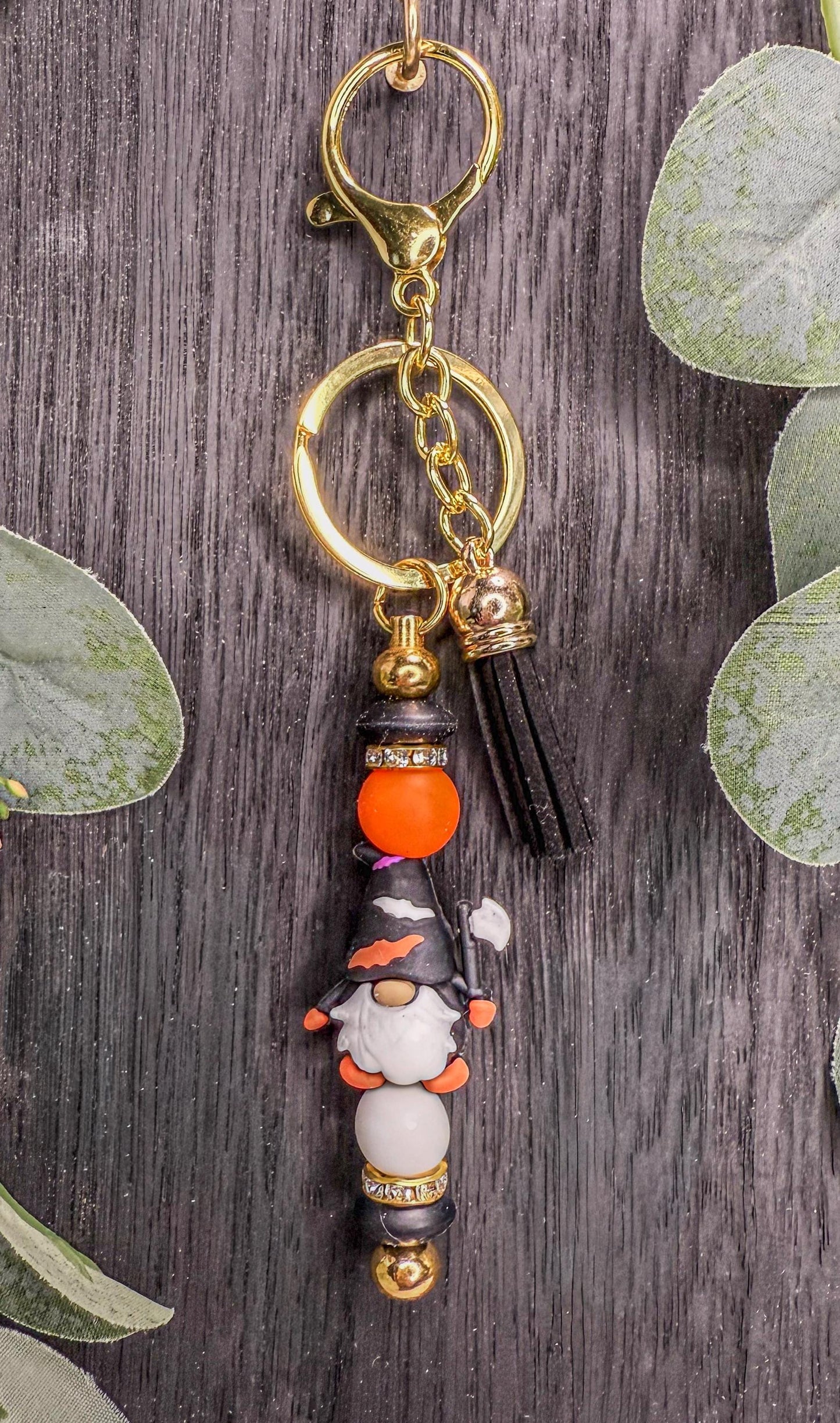 A keychain with a gnome-shaped charm and a tassel hanging from it, set against a wooden background with green leaves.