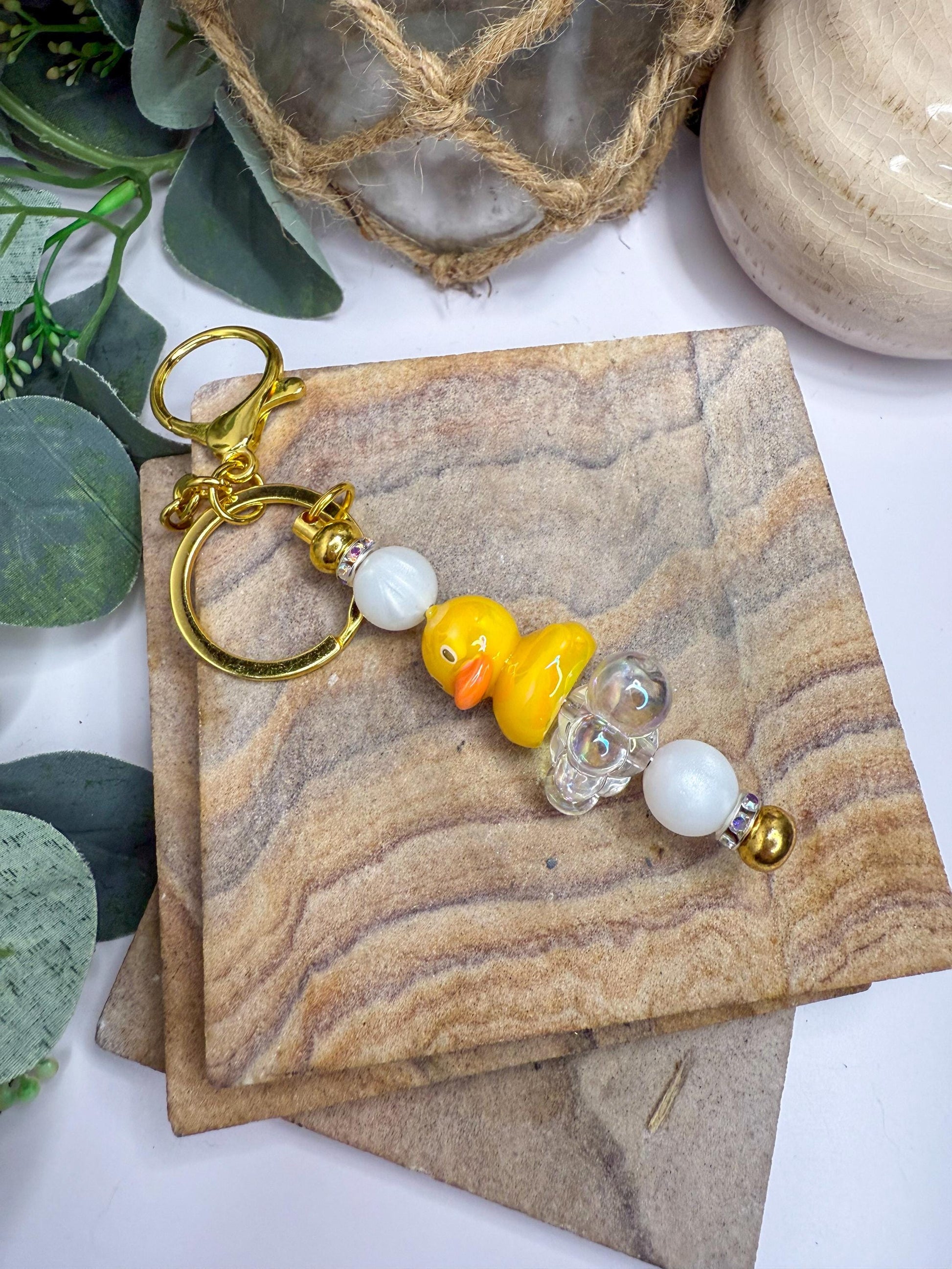 A keychain with a yellow duck charm and white beads is displayed on a wooden surface, surrounded by greenery.