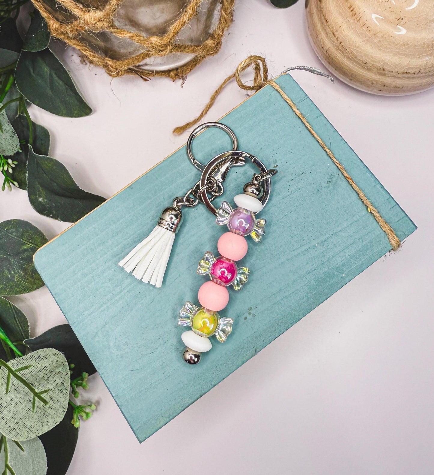 A keychain with colorful beads and a tassel is placed on a blue surface, surrounded by greenery.