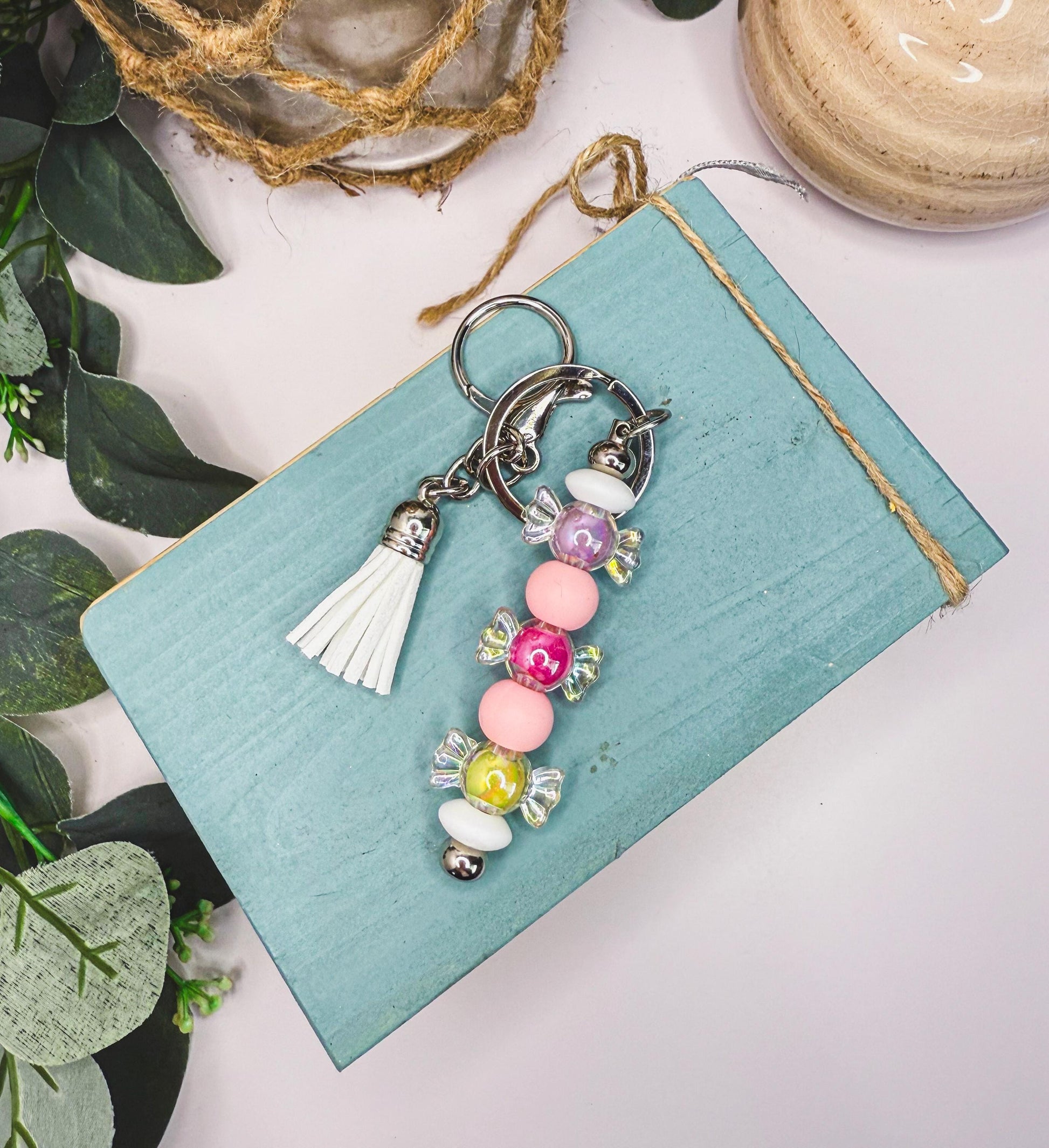 A keychain with colorful beads and a tassel is placed on a blue surface, surrounded by greenery.