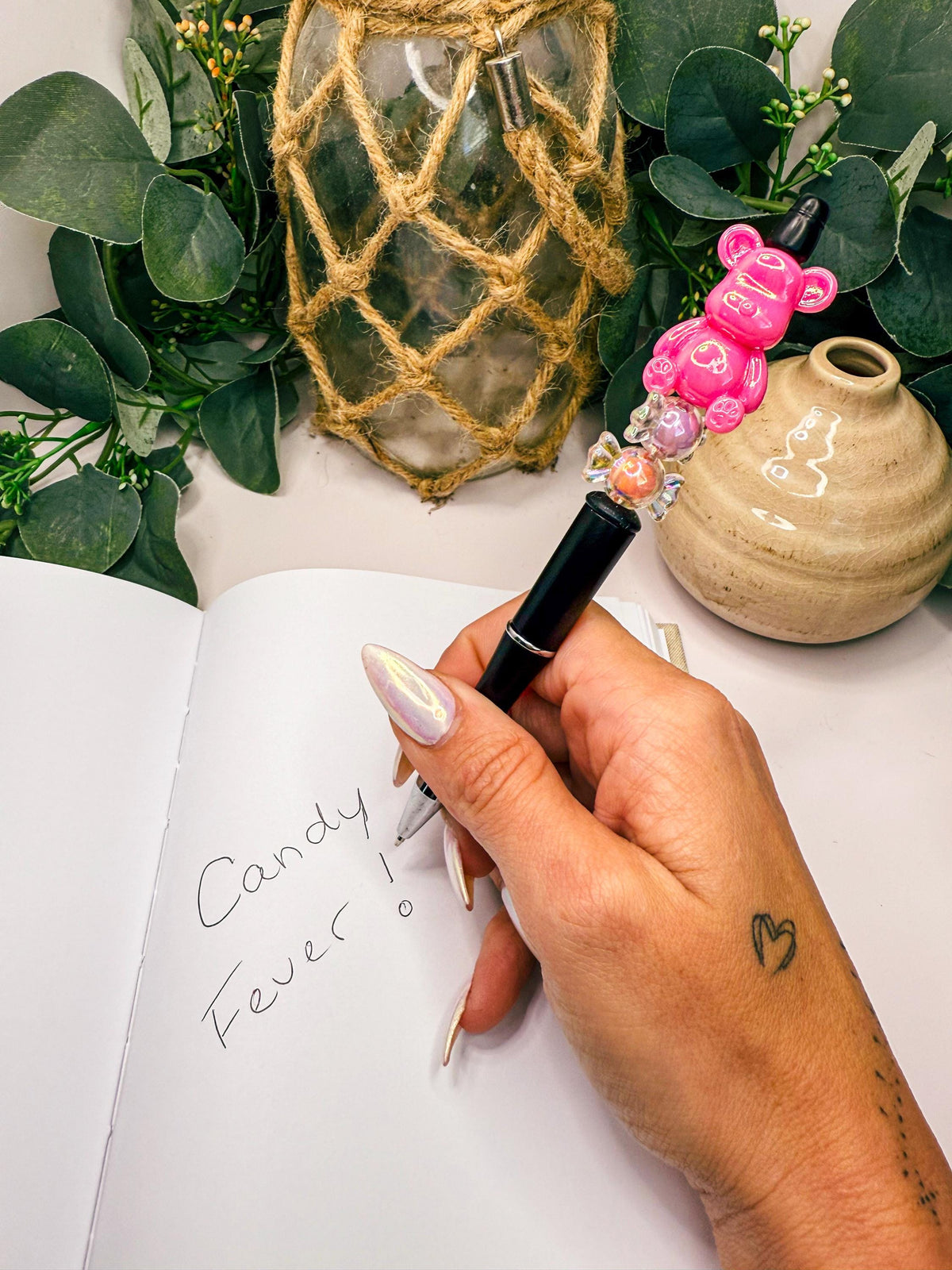 A hand holding a pen with a message written on a notebook, surrounded by a plant and a small ceramic object.