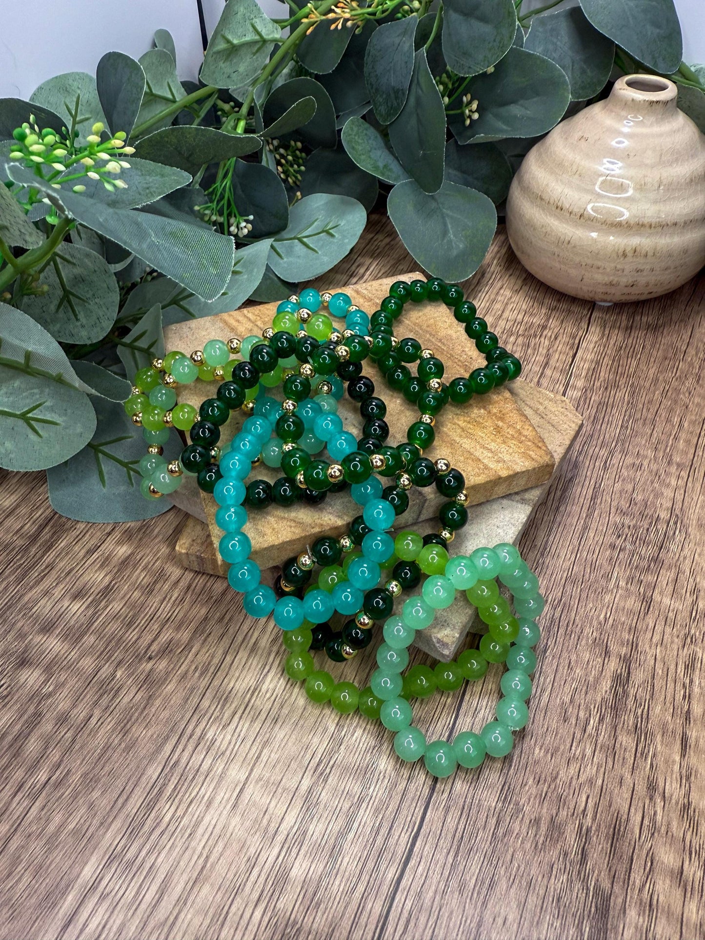 a collection of green and blue beaded bracelets arranged on a wooden surface, with a vase and greenery in the background.