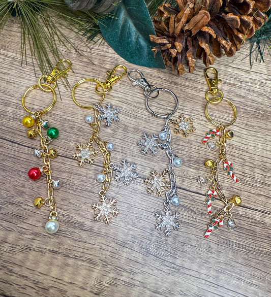 May include: Four festive keychains with gold, silver, and pearl accents. Each keychain features a different design: colorful bells, snowflake charms, and candy cane pendants. The keychains are displayed on a wooden surface with pine needles and a pine cone.