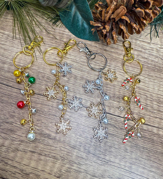 May include: Four festive keychains with gold, silver, and pearl accents. Each keychain features a different design: colorful bells, snowflake charms, and candy cane pendants. The keychains are displayed on a wooden surface with pine needles and a pine cone.