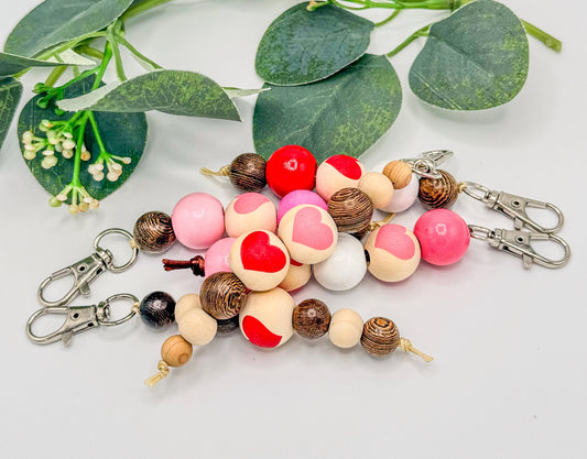 May include: A collection of beaded keychains with wooden beads in various colors, including pink, red, and brown. Some beads feature heart designs. Each keychain has a silver-colored clasp.