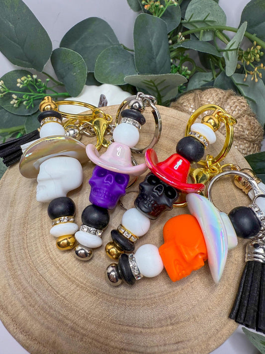 May include: A collection of keychains featuring skull charms with cowboy hats in various colors, including white, purple, red, and orange. The keychains have beaded accents and metallic key rings, with black tassels.