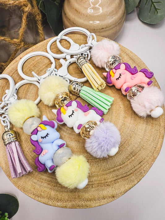 May include: Keychains with unicorn charms, fluffy pom-poms, and tassel accents. The keychains feature white metal rings and clasps. The unicorns are in various colors, including pink, blue, and white, with purple manes and yellow horns. The pom-poms are in shades of pink, yellow, and purple.
