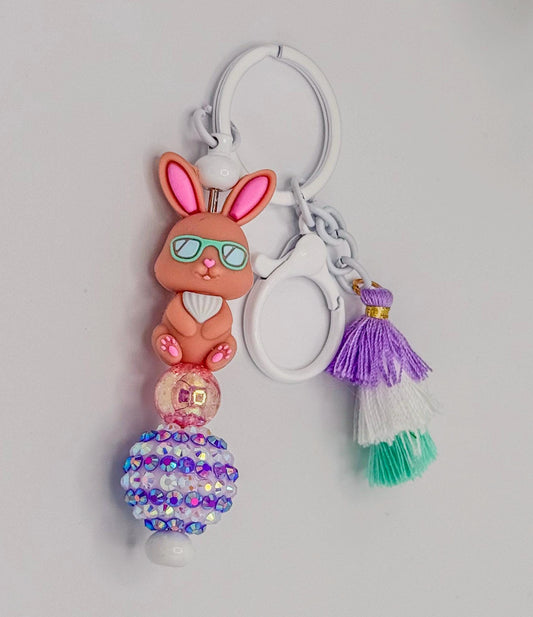 May include: A keychain featuring a brown bunny charm wearing sunglasses, with pink accents on the ears and feet. The keychain includes a beaded design with iridescent rhinestones and a purple, white, and green tassel.