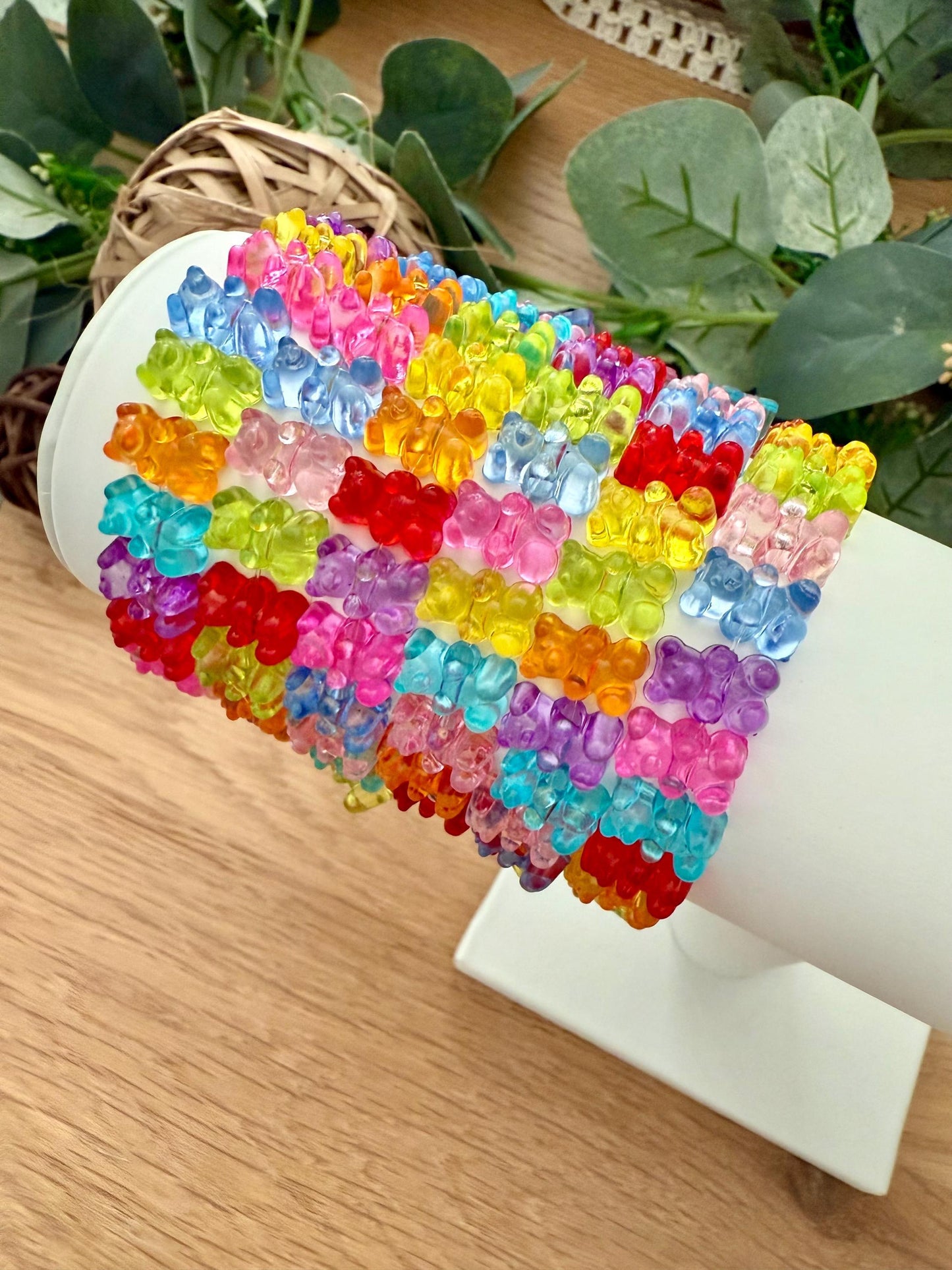 May include: A multi-strand bracelet featuring numerous small, translucent, colorful gummy bear-shaped charms in shades of red, orange, yellow, green, blue, and purple, arranged in a dense, repeating pattern around a white cylindrical display stand.